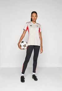 Žena v bielom a červenom futbalovom drese a čiernych nohaviciach, ktorá drží futbalovú loptu, stojí na jednoduchej svetložltej pozadí.