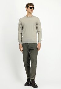 Mężczyzna w beżowym swetrze, oliwkowych spodniach, z podwiniętymi rękawami, ciemnych brązowych butach wizytowych i okularach przeciwsłonecznych, stojący na tle białego tła.