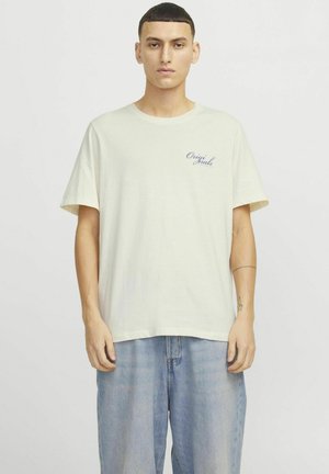 Camiseta de algodón beige claro con mangas cortas, que presenta un pequeño logo azul marino en el lado izquierdo del pecho. Combinada con unos vaqueros azules de corte holgado.