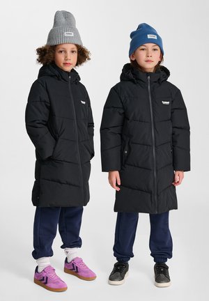 Svarta vadderade jackor med dragkedja, bärs över marinblå sweatpants. Den ena har en grå mössa, den andra en blå mössa. Skor: lila och svarta sneakers.