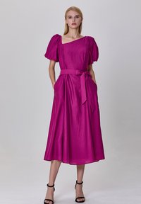 Robe fuchsia texturée avec manches bouffantes, design décolleté, lien à la taille et poches latérales. Silhouette évasée, longueur genou.