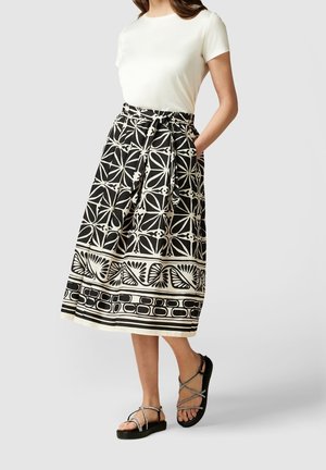 Femme portant un haut blanc à manches courtes associé à une jupe midi à motifs noir et blanc et des sandales noires à brides.