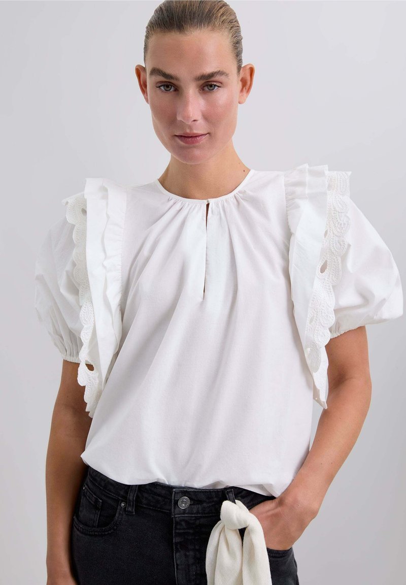 Weißes Blusenshirt mit puffärmel, Rüschenkanten und Spitzenbesatz. Besitzt einen Schlüssellochausschnitt und ein gerafftes Taillendesign. Getragen mit dunklen Jeans.