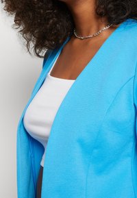 Turkos kavaj i stickad tyg, med djup V-ringning och öppen fram. Bärs över en vit, figurn ära tanktop. Silverhalsband.