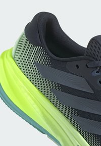 Schwarzer und neongrüner Sportschuh mit einem Mesh-Obermaterial, schlankem Design und drei dunkelgrauen Streifen an der Seite. Strukturiertes Profil.