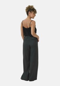 Donna con capelli ricci che indossa un top senza maniche nero e pantaloni a gamba larga scuri, in una posa di tre quarti vista da dietro.