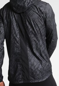 Chaqueta atlética negra con un patrón texturizado, que cuenta con capucha y paneles de ventilación en la parte posterior para una mejor transpirabilidad.