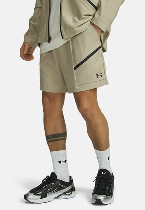 Man draagt beige Under Armour shorts en jas, witte sokken en zwarte en zilveren hardloopschoenen, staat met één hand in de zak.