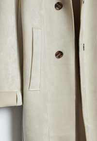 Gros plan d’un manteau en daim beige montrant une poche latérale et deux boutons ronds foncés sur l’ouverture avant.