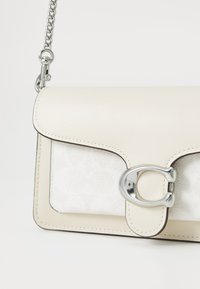 Beige läder crossbody-väska med en vit texturerad front, silverdetaljer och ett klaffförsedda stängning med en logotypaccent. Kedjesnodd ingår.