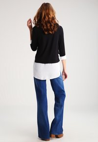 Svart tröja med trekvartslånga ärmar lager-på-lager över en vit skjorta, kombinerad med ljusblå utsvängda jeans. Outfiten har en avslappnad passform och en informell stil.