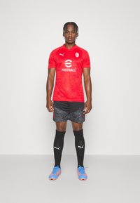 Maglia da calcio rossa a maniche corte, con un motivo a pois e il logo. Pantaloncini neri con motivi geometrici. Scarpette blu e calze nere.