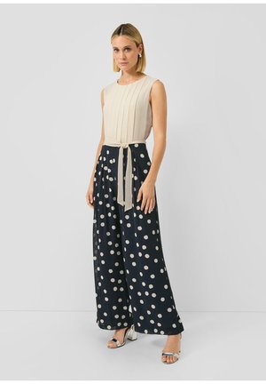 Kobieta w bezrękawniku w kolorze beżowym z wiązaniem w talii, zestawionym z szerokimi czarnymi spodniami w białe kropki oraz srebrnymi szpilkami.