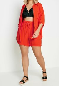 Czerwona koszula krótki rękaw, rozpięta na guziki, w zestawie z czarnym topem kąpielowym z otworem w kształcie dziurki. Czerwone szorty z elastycznym pasem i sznurkiem. Czarne sandały.