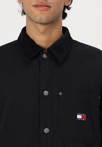 Svart skjorta i bomull med ett kordfloret krage, försedd med silverknappar och en vänster bröstficka med en flerfärgad logotyp.