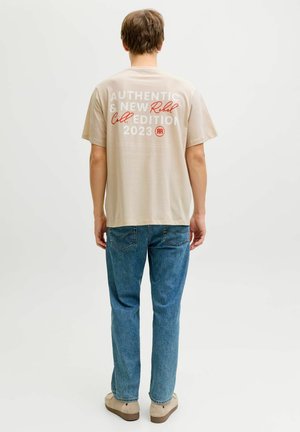 Giovane uomo in piedi, indossa una T-shirt beige con la scritta "Authentic & New Rebel Collection 2023", jeans blu e scarpe beige, di spalle.