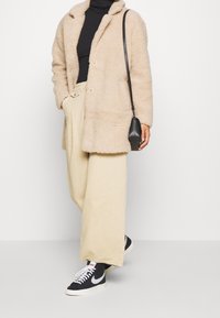 Beige, fyldig, oversized frakke med en notch krave, parret med en sort turtleneck, beige bukser og sorte sneakers med hvide detaljer.