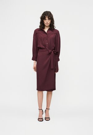 Vrouw met krullend haar, gekleed in een bordeauxrode jurk met lange mouwen en knopen, met een tailleceintuur en zwarte, open hakken met bandjes, staand tegen een witte achtergrond.