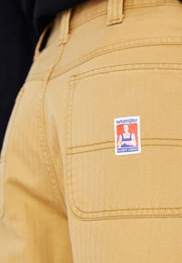 Texturált felületű bézs nadrág, látható Wrangler címke folttal a hátsó zsebben és hagyományos zsebvarrással.