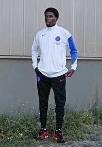 Hvid trackjakke med blå ærmer, prydet med et lilla cirkulært logo. Sorte bukser med rødt Nike-logo, parret med rød- og svart sneakers.