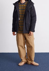 Schwarzer gepolsterter Parka mit Kapuze und mehreren Taschen, darüber ein grün gestreiftes Hemd. Hellbraune Cargohose und beige Sneakers.