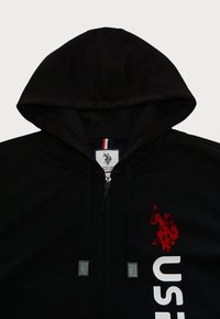 Hanorac negru cu fermoar, cu un text alb mare "US", emblema roșie de polo pe față și fermoar cu contrast, având o glugă căptușită.