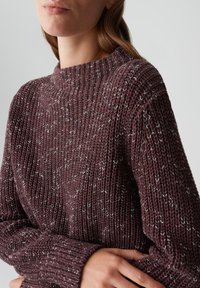 Eine Frau trägt einen grob gestrickten, kastanienbraunen Pullover mit langen Ärmeln und Rippendetails, die Arme an der Taille verschränkt, vor neutralem Hintergrund.