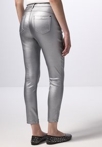 Stříbrné metalické slim-fit kalhoty s zadními kapsami, hladkým povrchem a zkrácenou délkou. Nosené s plochými černými botami s hroty.
