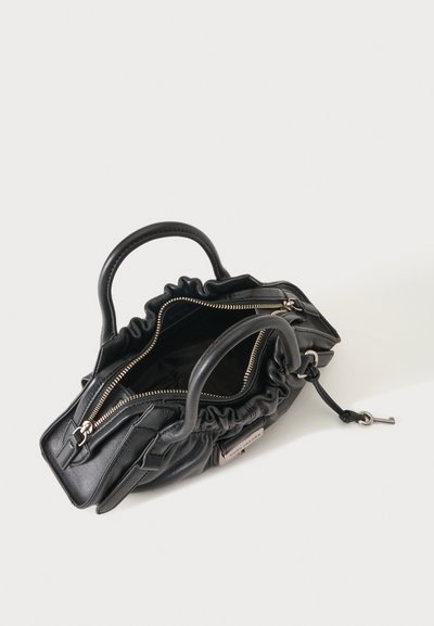 Sac à main en cuir noir avec deux poignées, fermeture éclair ouverte sur le dessus et clé suspendue à un cordon sur un fond clair uni.