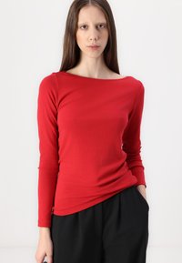 Rdeča dolgosleeve majica iz mehke tkanine, z načrtom v obliki čolna in prilegajočo se zasnovo. Združena s širokimi črnimi hlačami.