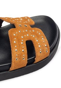 Sandal in suede marrone con dettagli borchiati, caratterizzato da un design con ampia cinghia e una suola nera testurizzata, che enfatizza comfort e stile.