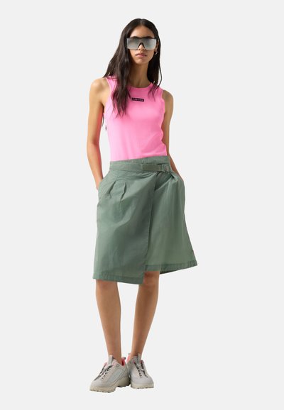 Frau trägt ärmelloses pinkes Oberteil, grünen Wickelrock mit Gürtel, graue Sneakers und große verspiegelte Sonnenbrille, steht mit den Händen in den Taschen.