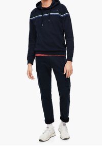 Felpa navy con cappuccio e lacci, decorata con strisce bianche, verdi e blu; abbinata a pantaloni cargo scuri e scarpe sportive bianche.