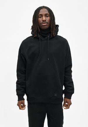 Mann mit Dreadlocks, der einen schwarzen Hoodie und schwarze Hosen trägt, steht vor einem einfarbigen weißen Hintergrund.