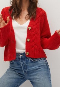 Cardigan rouge en tricot avec un motif torsadé texturé, doté de boutons dorés, de poignets côtelés et d'une coupe courte, assorti à un jean bleu clair.