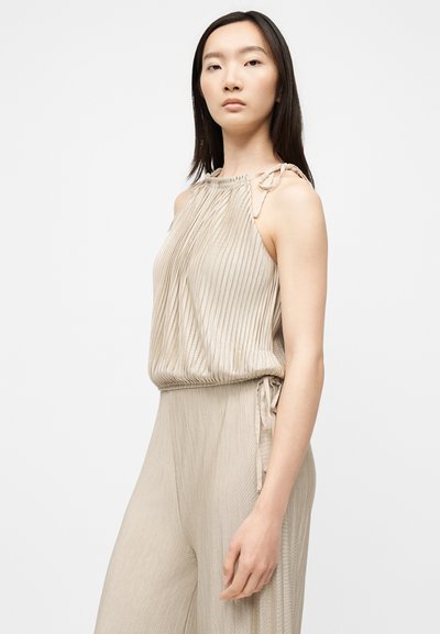 Femme aux cheveux raides et foncés portant un haut plissé beige sans manches avec bretelles à nouer et un pantalon large plissé assorti, sur fond blanc.