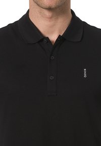 Polo noir en coton ; dotée d'un col traditionnel, de trois boutons et d'une broderie blanche "HUGO" sur la poitrine gauche.