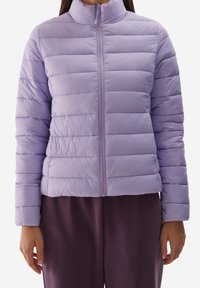 Giacca a vento viola chiaro con colletto alto, zip frontale e texture trapuntata. Indossata sopra pantaloni viola scuro.