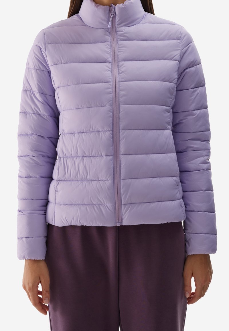 Giacca a vento viola chiaro con colletto alto, zip frontale e texture trapuntata. Indossata sopra pantaloni viola scuro.