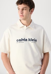 Bijela pamučna polo majica s preklopljenim ovratnikom. Na prednjoj strani nalazi se natpis u tamnoplavoj boji: „calvin klein“ i „West 39th Street, New York City.“