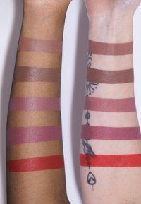 Échantillons de teintes à lèvres sur deux bras : différentes nuances de nude, de rose et un rouge vibrant. Un bras a un tatouage ; les teintes de peau varient.