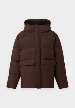 Jeune homme debout les mains dans les poches portant une doudoune marron foncé, une chemise bleu marine, un pantalon beige à jambes larges et des chaussures foncées.