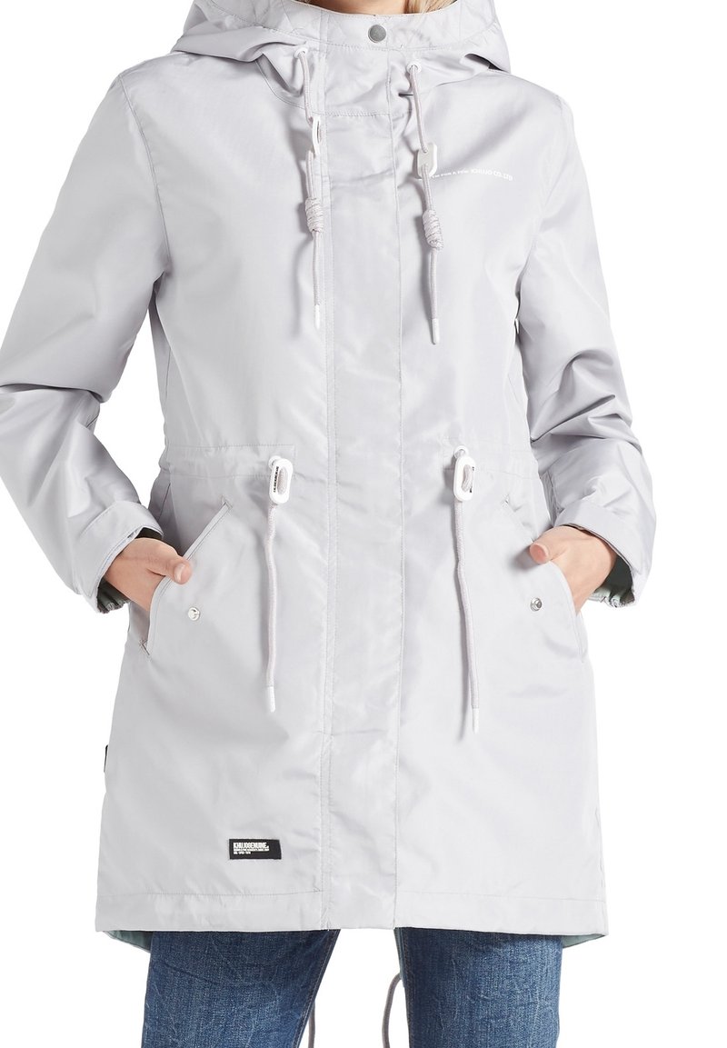 Světle šedý voděodolný parka s kapucí na šňůrku, bočními kapsami, elastickými manžetami a o něco delším zadním lemem. Obsahuje minimální branding.