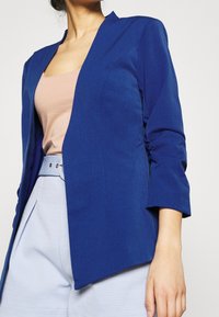 Blazer bleu à texture lisse, à ouverture frontale et manches trois-quarts. Porté sur un haut rose pâle et un pantalon bleu clair à motifs.
