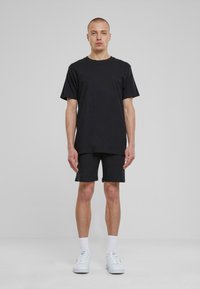T-shirt en coton noir à manches courtes associé à un short foncé. Le modèle porte des baskets blanches et des chaussettes blanches jusqu'aux chevilles. Fond neutre.