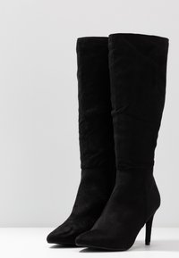 Un par de botas altas de ante negro con punteras puntiagudas y tacones delgados y altos, de pie sobre una superficie blanca frente a un fondo liso.