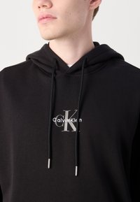 Sudadera con capucha negra hecha de una tela suave, que presenta un logo en la parte frontal con el texto "Calvin Klein" y detalles en plateado en los cordones.