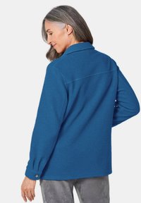 Blauwe fleeceshirt met een kraag en knoopmanchetten, met een relaxte pasvorm en een gladde textuur, weergegeven vanuit de achterkant.