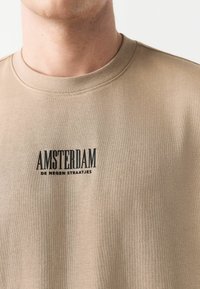 Camiseta de algodón beige con textura acanalada y cuello redondo. Presenta texto impreso en negro: "AMSTERDAM DE NEGEN STRAATJES" en la parte frontal.