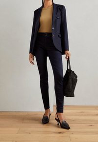 Blazer azul marino sobre una blusa ajustada color arena, combinado con pantalones de mezclilla oscuros y mules negros de tacón. Accesorio: bolso negro texturizado con cordón.
